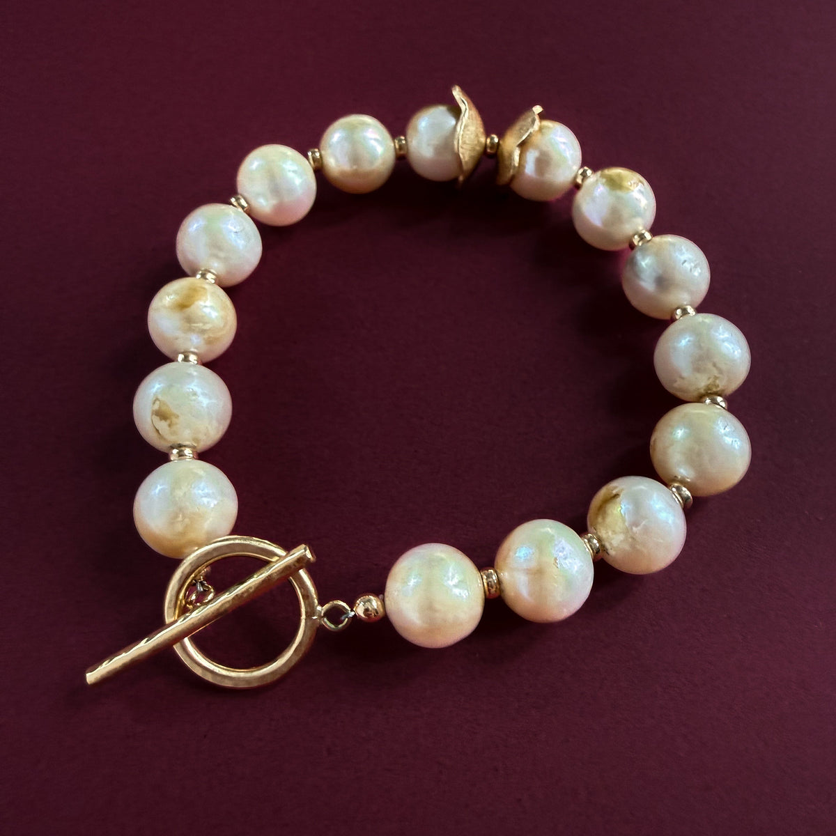 A pearl bracelet made of round, lustrous off-white pearls with gold-filled spacer beads and a gold-filled toggle clasp, displayed on a dark burgundy background.