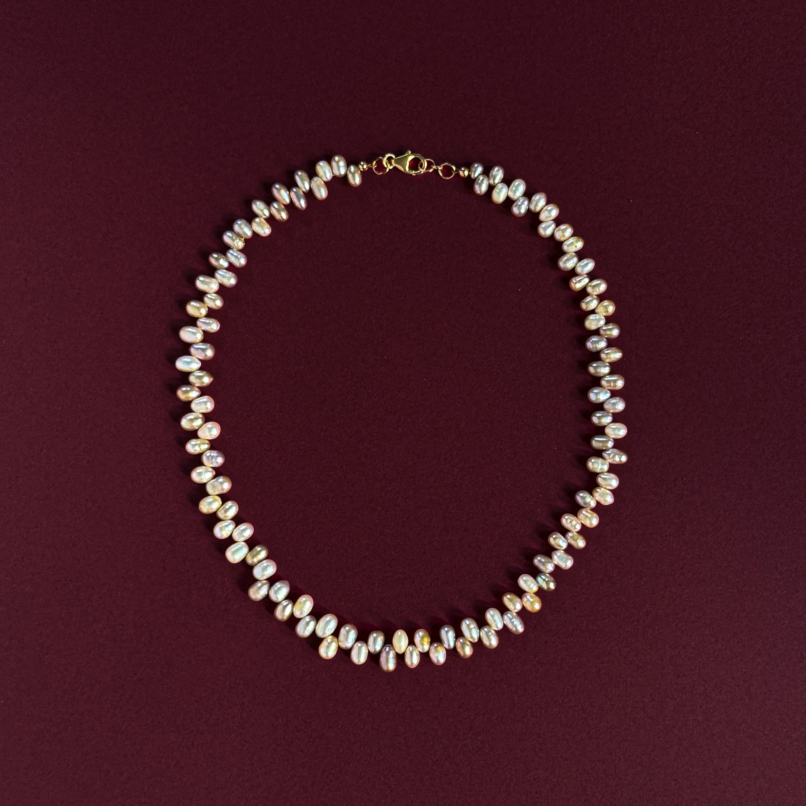 A delicate necklace made of small iridescent baroque pearls on a gold-tone chain, arranged in a circle on a dark burgundy background.