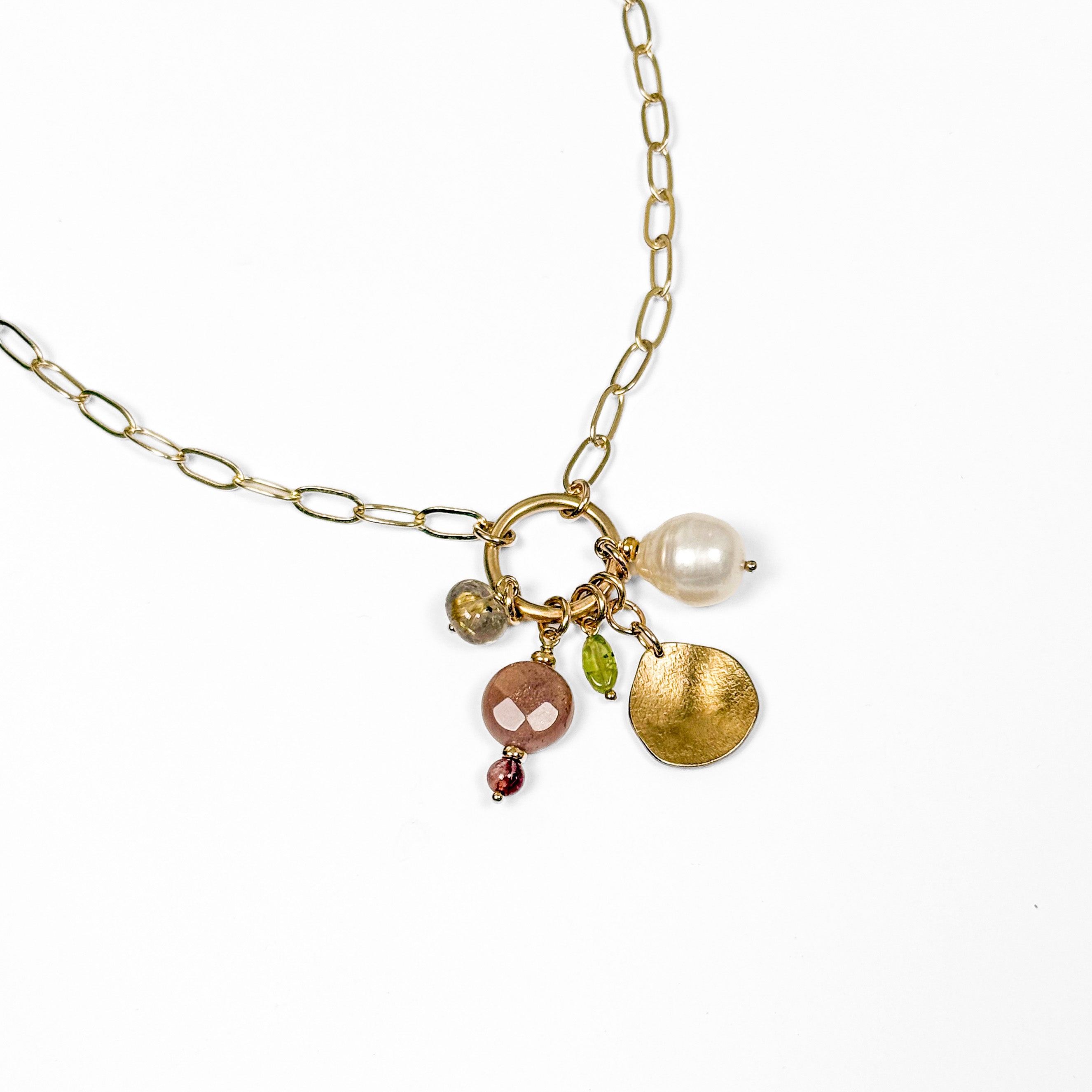A gold-filled chain necklace with a ring clasp holding multiple charms, including a white pearl, a textured gold disc, a pink stone, a green peridot bead, and a small red bead, shown on a white background.