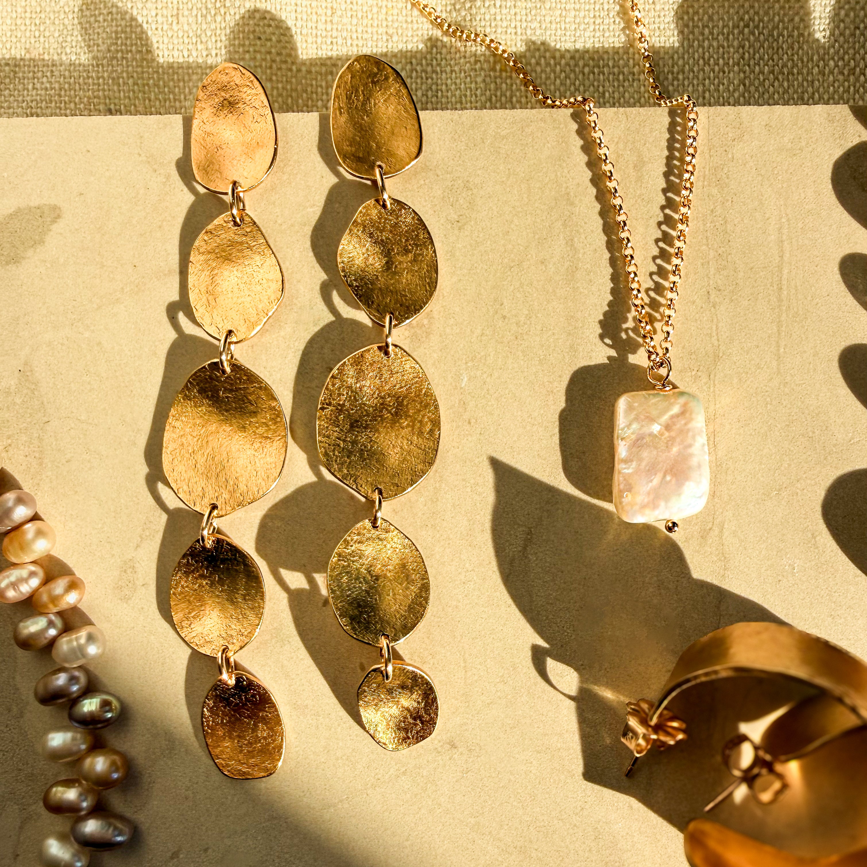 Gold-toned jewelry arranged on a beige surface in warm sunlight, including a pair of dangling pebble-shaped earrings, a necklace with a rectangular pearl-like pendant, and partial pearl strands and hoop earrings.