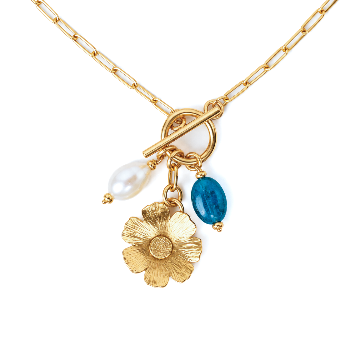 Gold paperclip chain necklace with a toggle clasp and three dangling charms: a white pearl bead, a textured gold flower, and a blue oval gemstone, shown on a white background.