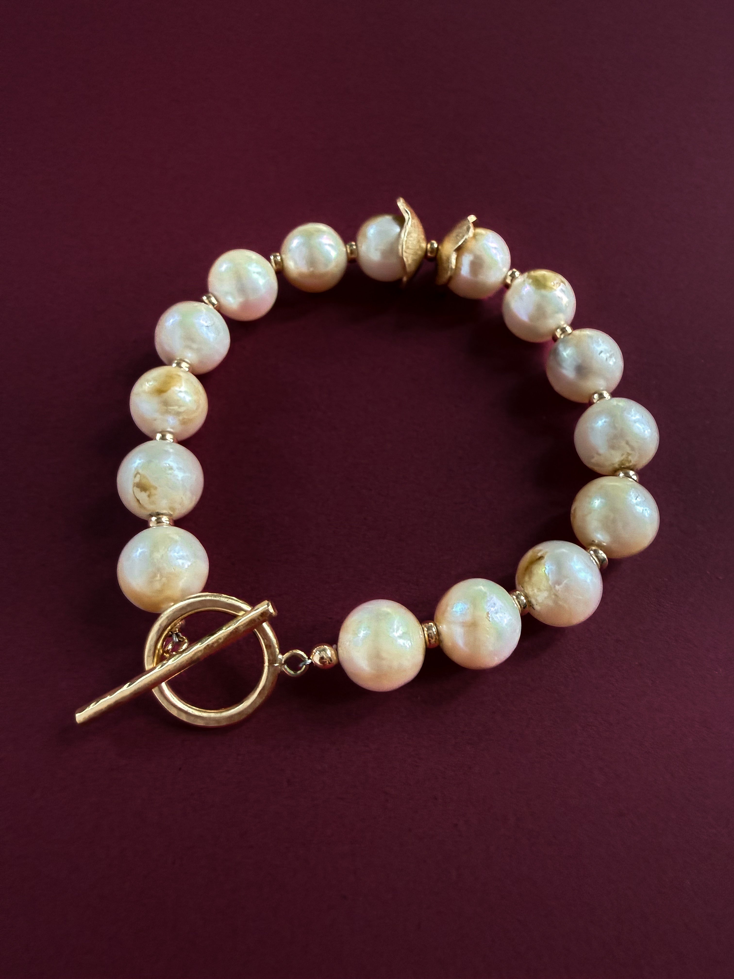 A pearl bracelet made of round, lustrous off-white pearls with gold-filled spacer beads and a gold-filled toggle clasp, displayed on a dark burgundy background.