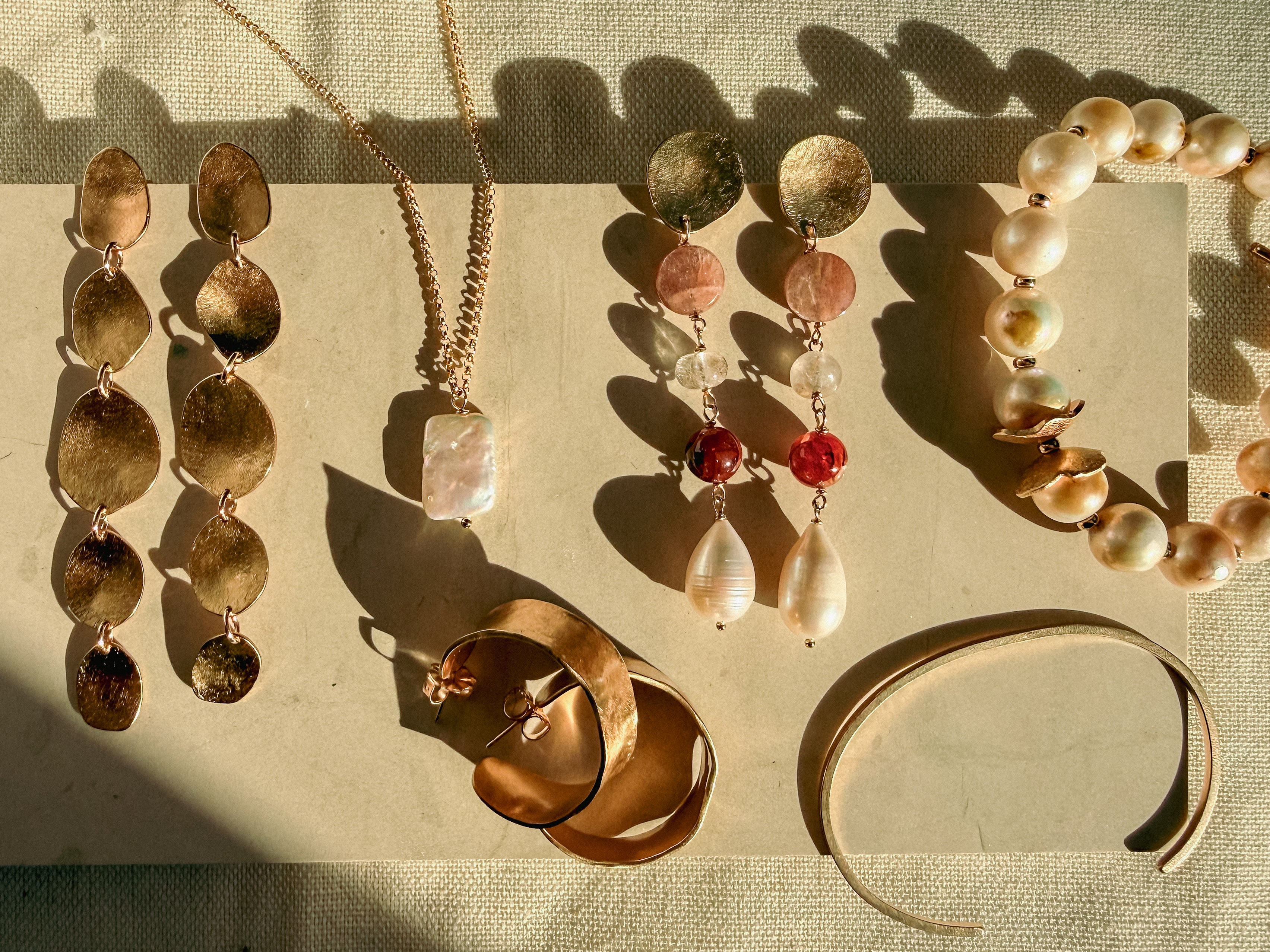 A styled flat-lay of multiple jewelry pieces on a beige fabric background with strong sunlight and shadows. The collection includes gold-toned disc drop earrings, a gold pendant necklace with a white stone, a pair of dangling earrings with pink and red beads and white teardrop pearls, a pearl beaded bracelet, a gold cuff bracelet, and a gold headband.