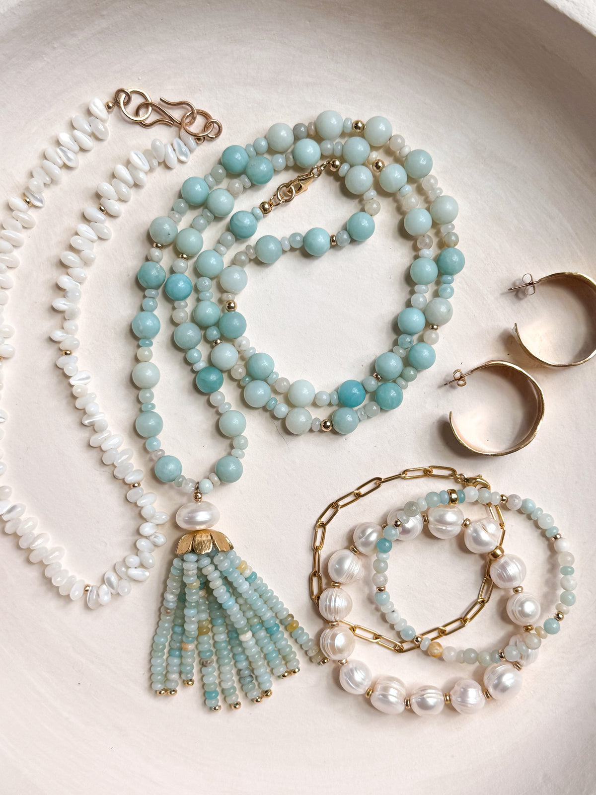 A jewelry set arranged in a shallow round dish, featuring a long necklace with white shell beads and a tassel, a layered necklace with light blue beads, gold hoop earrings, and multiple pearl and bead bracelets.