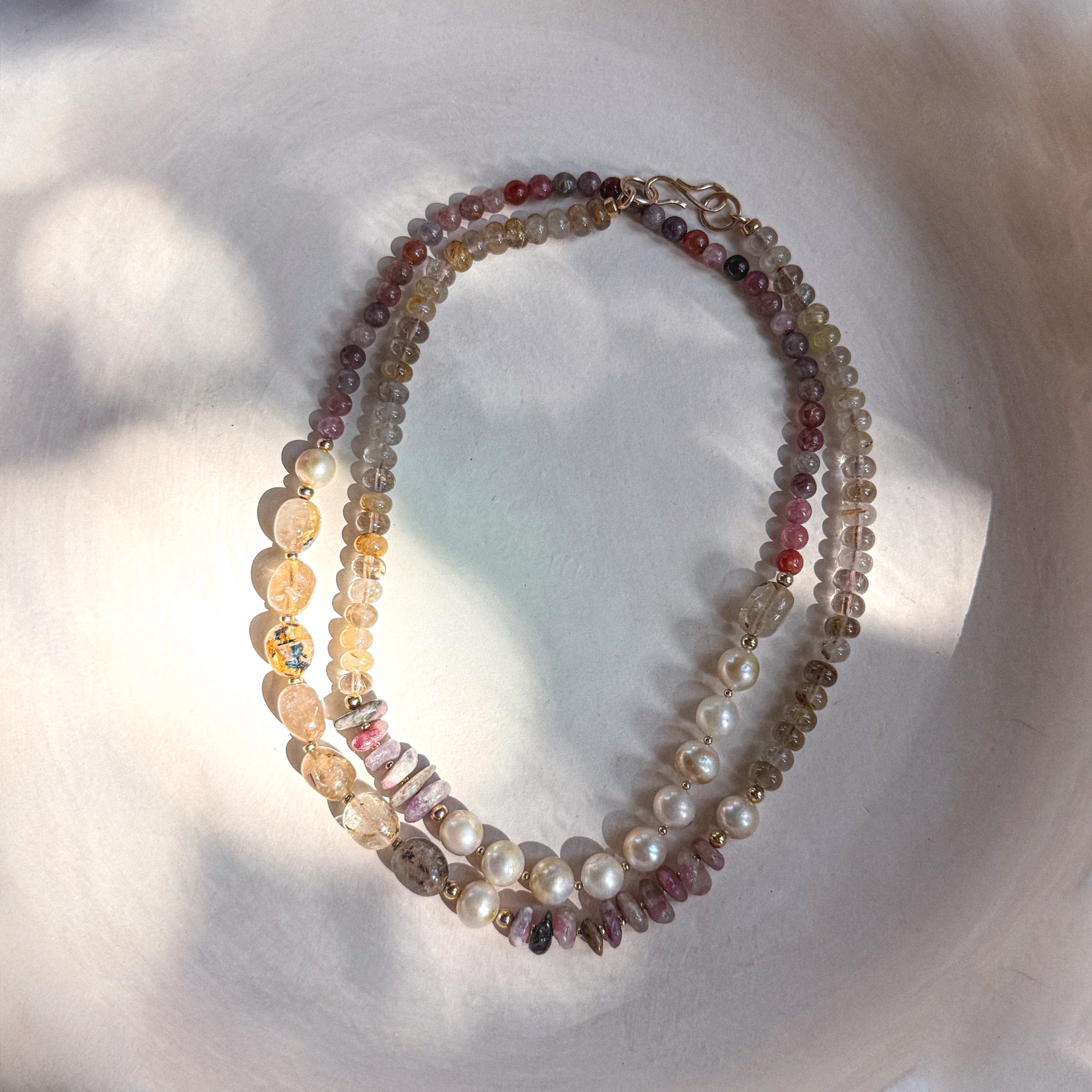 A multi-strand beaded necklace with mixed gemstone beads, white pearls, and gold-toned accents, arranged in an oval on a white textured surface with soft shadows.