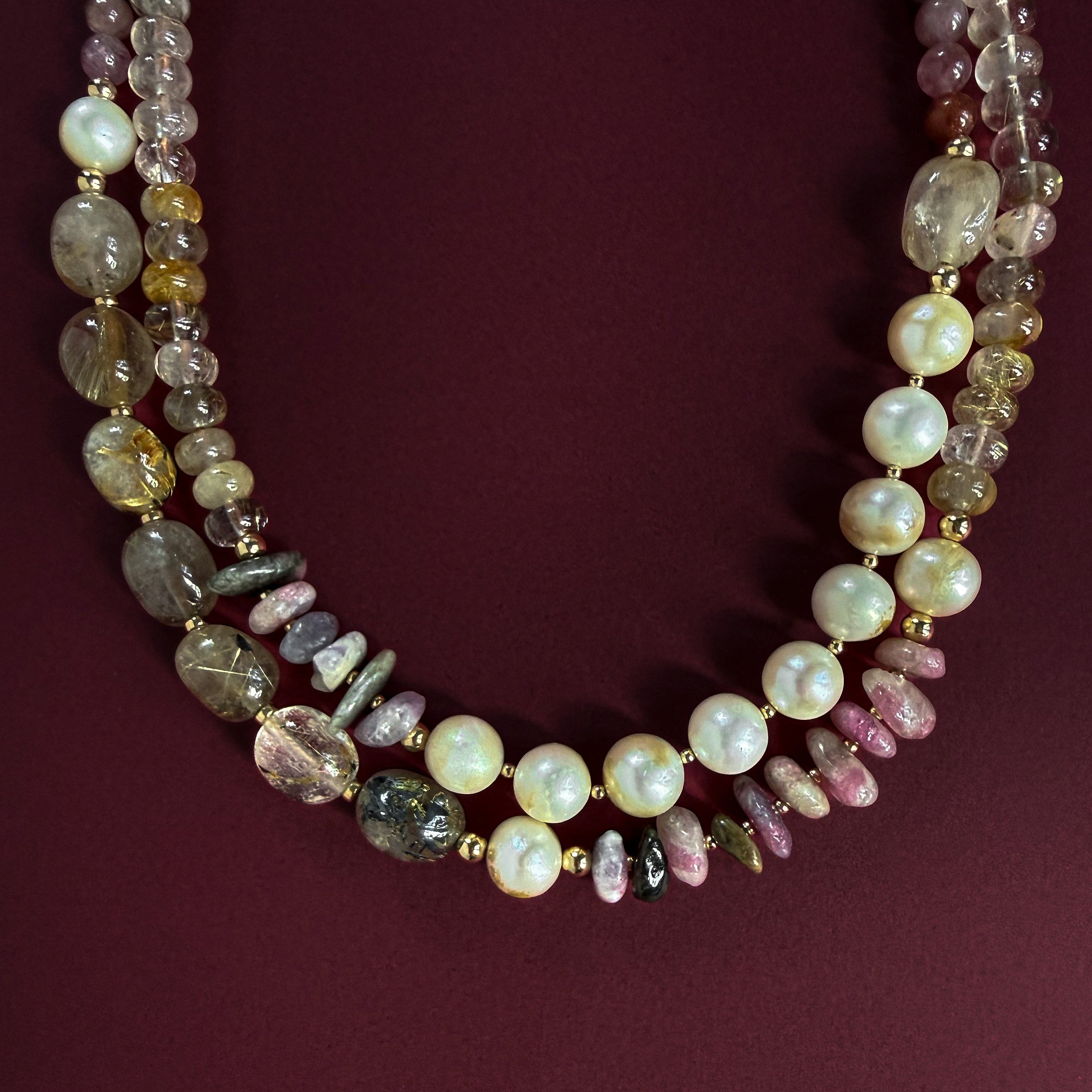 A multi-strand beaded necklace featuring white pearls, rutilated quartz, pink tourmaline, and polished gold-filled beads, displayed on a dark burgundy background.