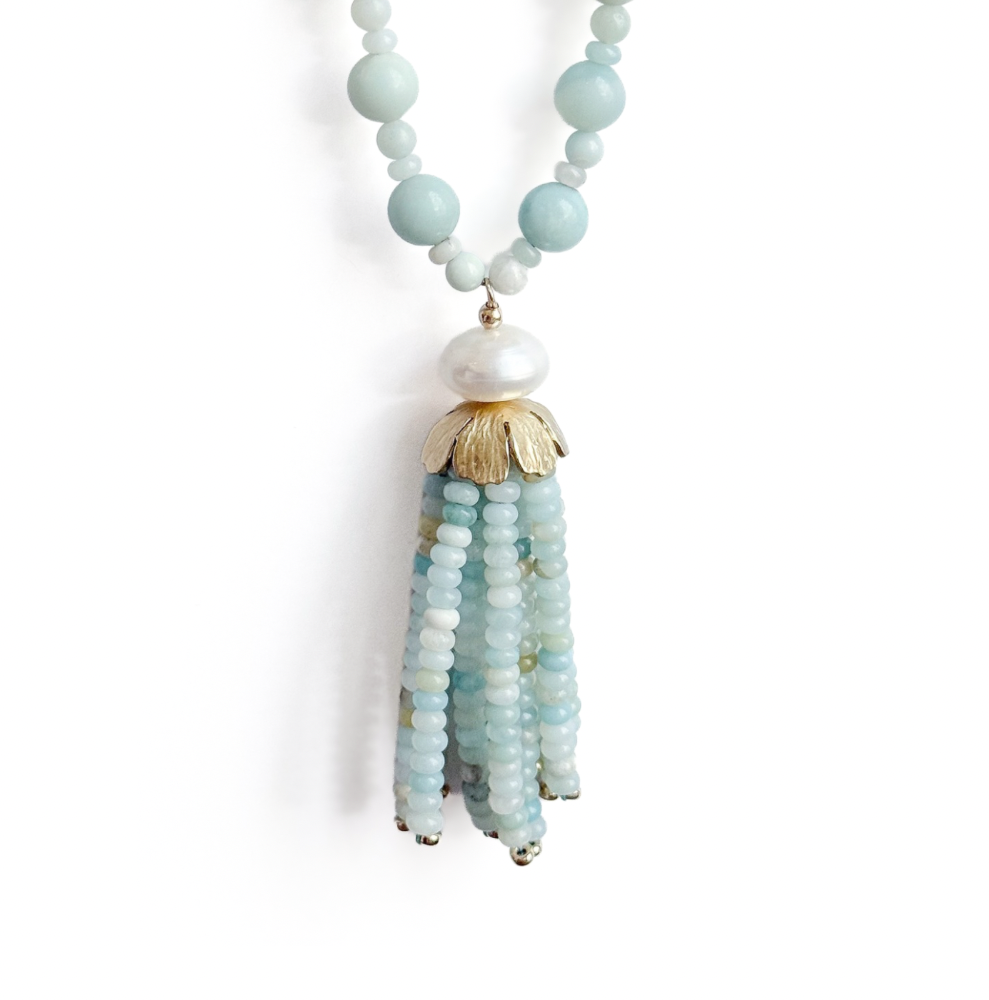 A beaded tassel necklace featuring light blue and white beads, a white pearl, and gold-toned metal accents, displayed on a clean white background.
